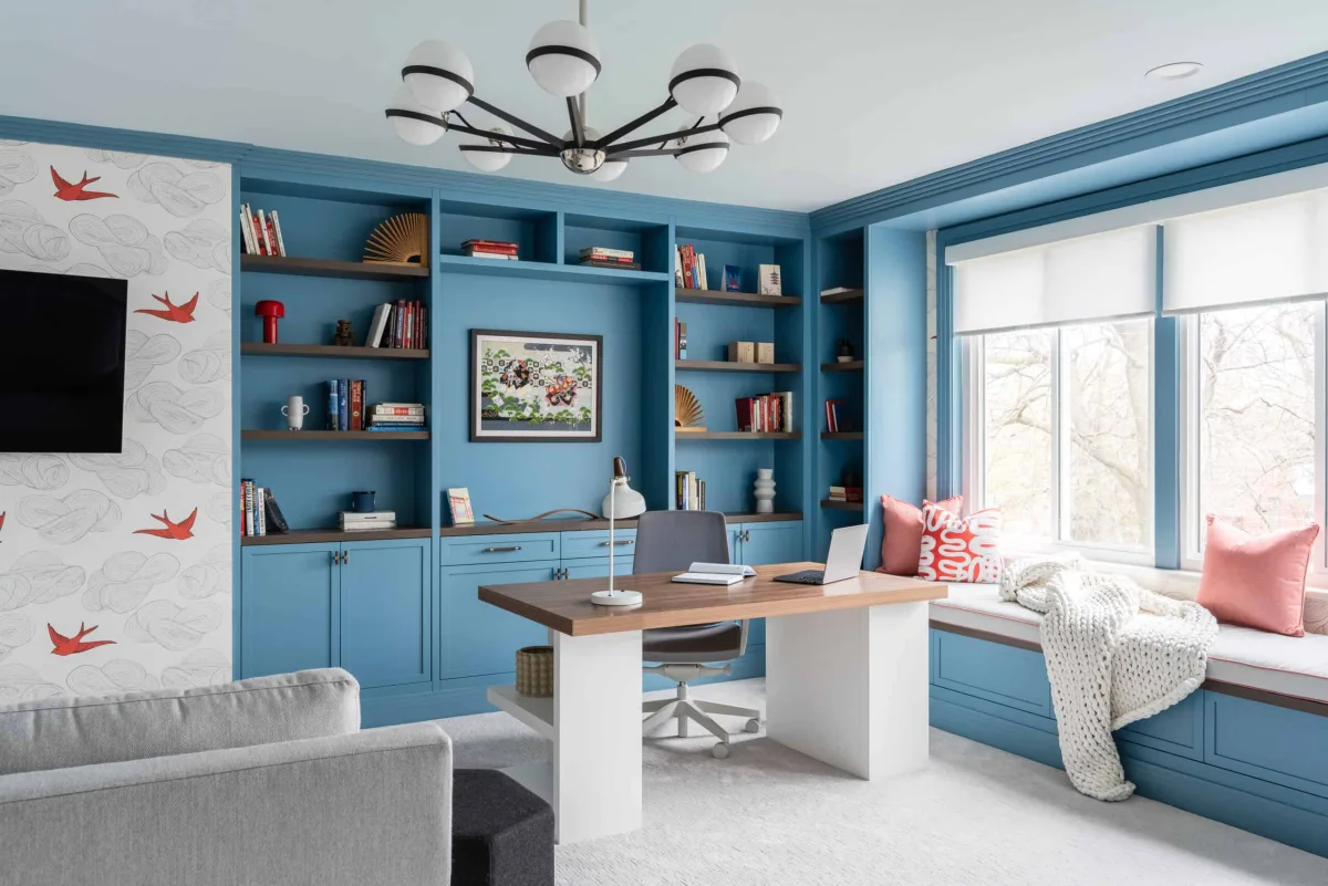 Home office with blue shelving, a wood desk, patterned curtains and a cushioned window seat.