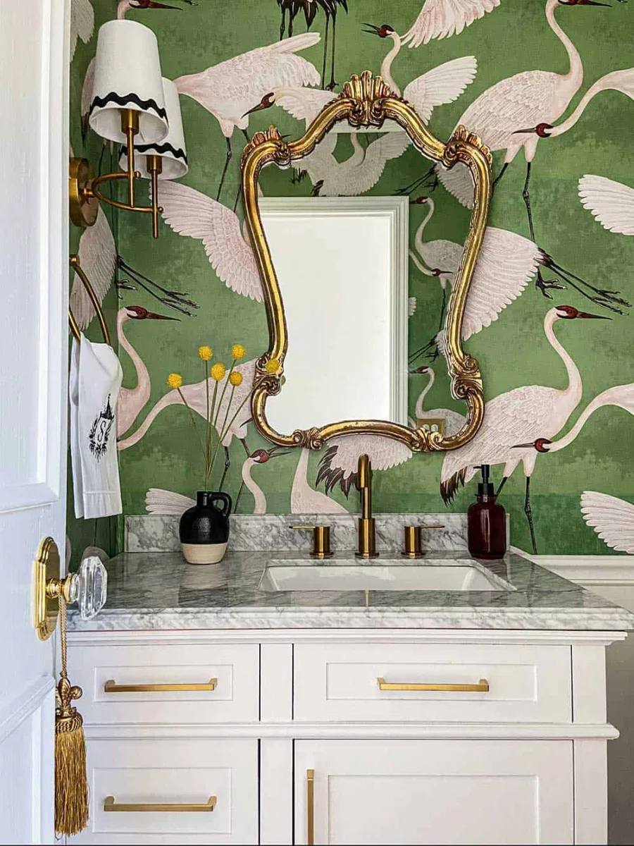 Green powder room with heron wallpaper, a gilded mirror and a white marble-topped vanity.